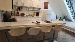 Une cuisine moderne avec des meubles blancs, une cuisinière à gaz, une machine à café et un réfrigérateur. On y trouve trois tabourets de bar et un petit îlot central avec une assiette et une serviette.