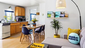 A modern kitchen with wooden cabinets, a dining area with a table and chairs, a sofa with a 'make waves' pillow, and a bookshelf with travel books. - Seascape Motel & Cottages (Belfast)