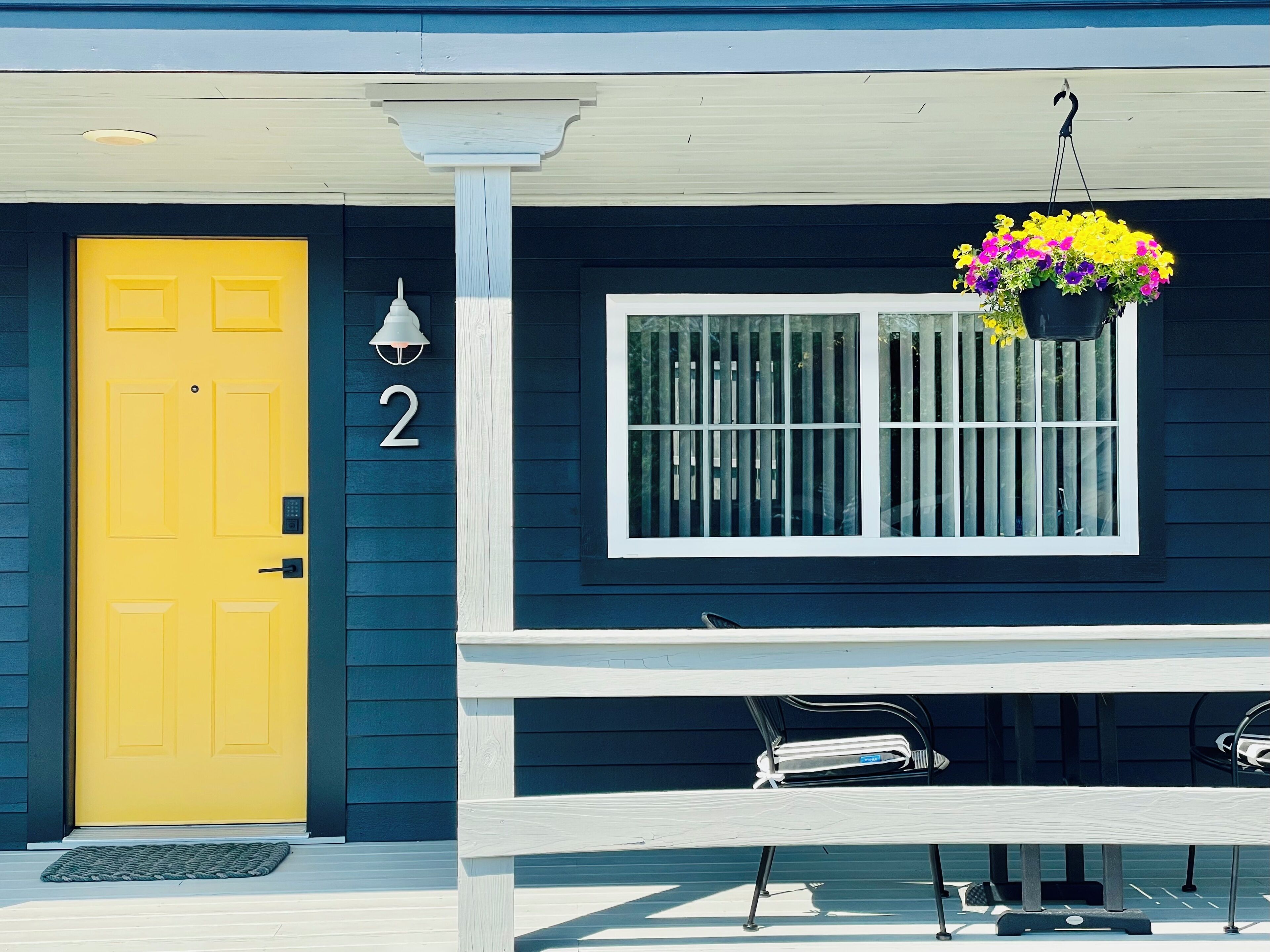A house with a yellow door, a number '2' on the wall, a window with bars, and a hanging flower pot.