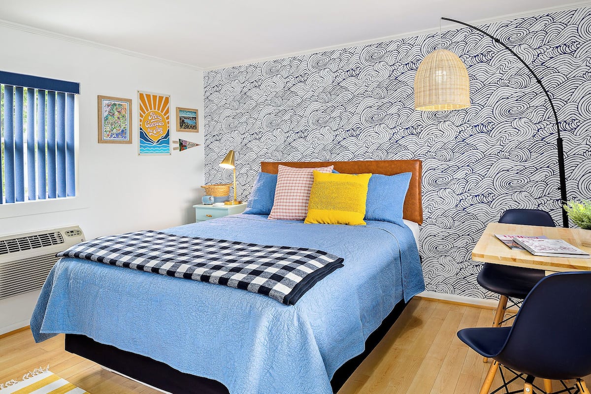 A bedroom with a bed, a desk, and a chair. There is a window with blinds, a wall with a wavy pattern, and a lamp on the desk.