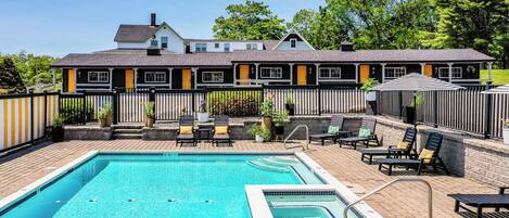 A swimming pool with lounge chairs and a covered seating area, surrounded by a fence and a building with numbered rooms.