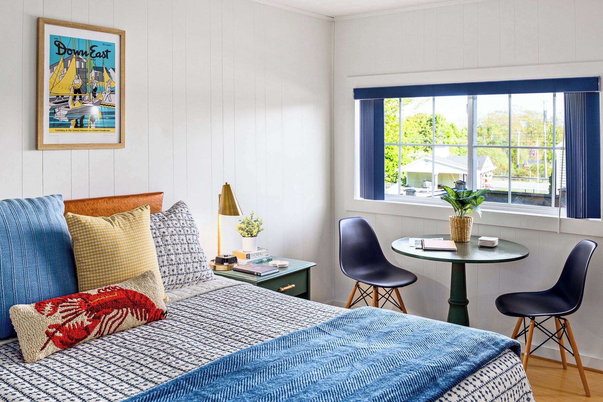 Ein Zimmer mit einem Bett, zwei Stühlen, einem kleinen Tisch, einem Fenster mit Vorhängen, einer Pflanze und einem gerahmten Bild an der Wand.