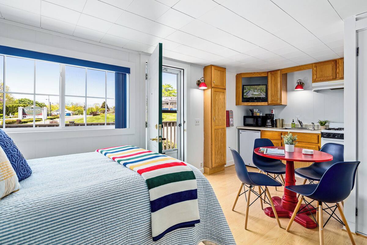 A room with a bed, a dining table with chairs, a kitchen area, and a view of the outside.