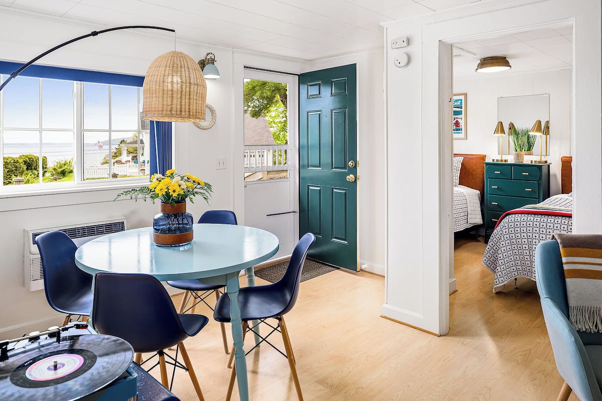 A room with a round table, chairs, a window with a view, a door, and a bed with a headboard.