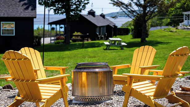 A patio with three yellow Adirondack chairs and a metal fire pit. In the background, there is a wooden cabin, a picnic table, and a view of a body of water. - Seascape Motel & Cottages (Belfast)