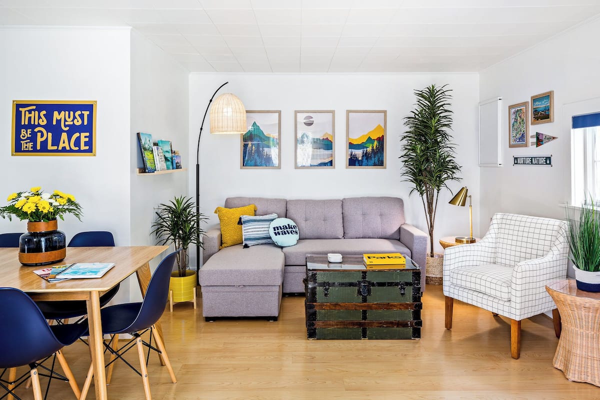 A modern living room with a sofa, a dining table with chairs, a coffee table, and various decorative items including a sign, framed pictures, and plants.