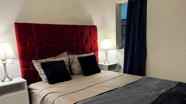 Een hotelkamer met een rood, gestuufd hoofdeinde, twee bedlampen, een bed met een beige- en zwart gestreept deken en een donkerblauw gordijn.