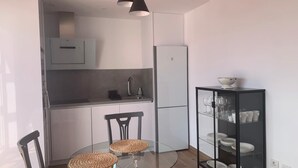 Una cocina compacta con una mesa redonda de vidrio, dos sillas, un armario negro con vajilla y una lámpara colgante moderna.