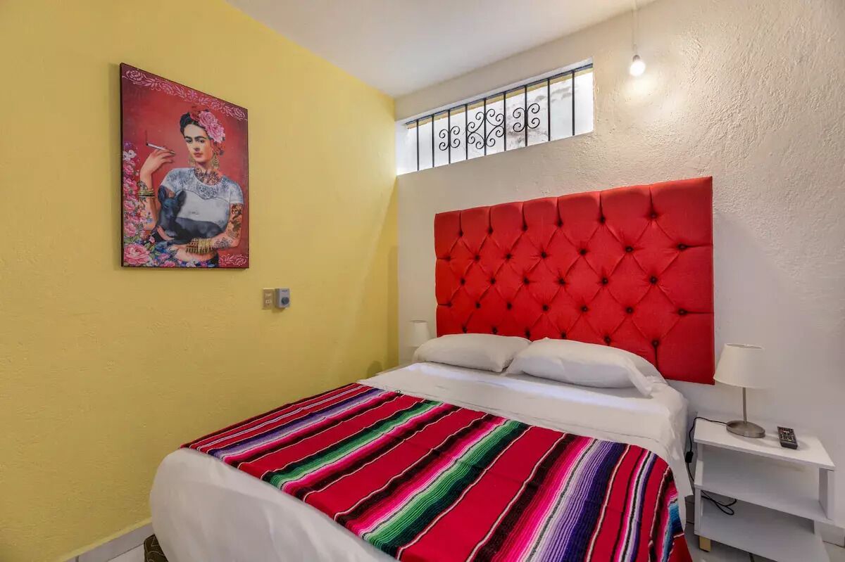 Un dormitorio con cabecera tapizada en rojo, colcha a rayas coloridas, mesita de noche blanca con lámpara y un cuadro en la pared.