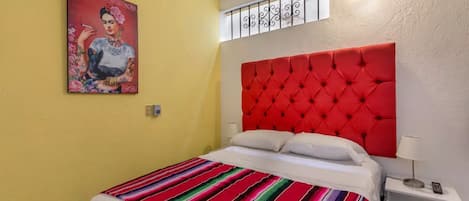 A bedroom with a red tufted headboard, a colorful striped bedspread, a white bedside table with a lamp, and a painting on the wall.