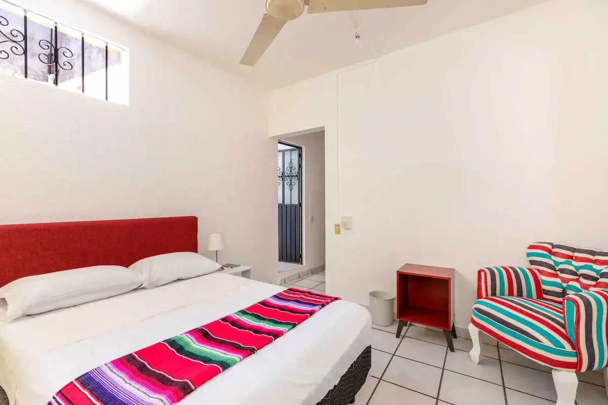 Un dormitorio con cama, cabecera roja, una silla con estampado colorido, una mesita y una puerta que da a otra habitación.