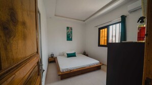 A bedroom with a wooden bed frame, white bedding, a green pillow, a nightstand, a wall-mounted air conditioner, and a window with curtains.