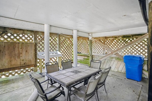 An outdoor patio with a table and chairs, a blue trash bin, and a wooden fence.