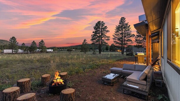 An outdoor seating area with a fire pit, a sofa, and a table, surrounded by trees and a sunset view. - Grand Canyon Brand New Home with AC. Near Golf Course, Lake & Bearizona (Williams)