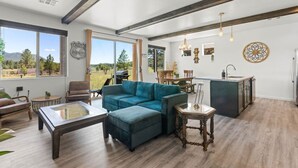 A spacious living area with a large sofa, a coffee table, and a dining table. There are large windows offering a view of the outdoors. - Grand Canyon Brand New Home with AC. Near Golf Course, Lake & Bearizona (Williams)