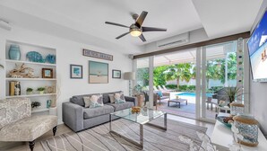 A modern living room with a grey sofa, a glass coffee table, and a ceiling fan. There are shelves with decorative items and a sign that reads "Beach House". - Steps from Beach | Heated Pool | Gulf Stream Key | RESlDENCES (Fort Lauderdale)
