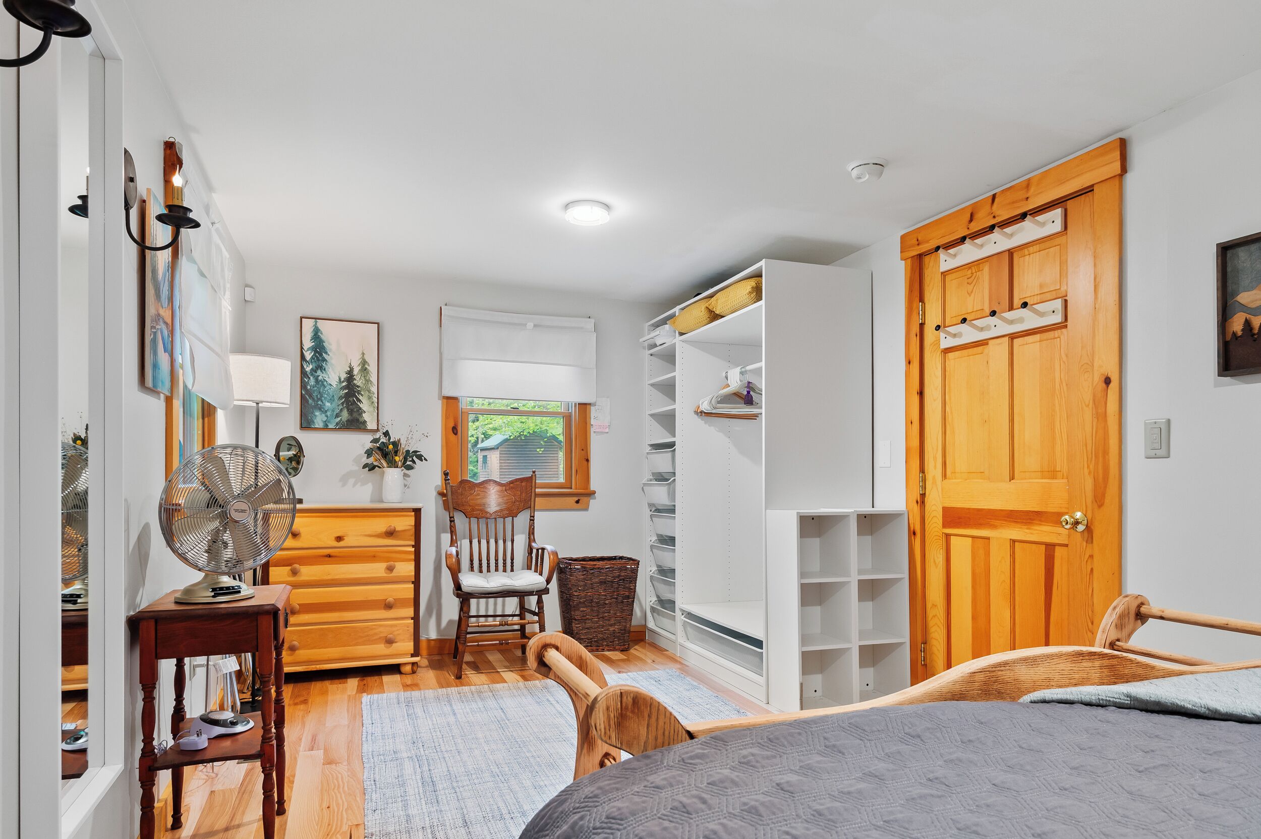 A bedroom with a bed, a wooden dresser, a chair, a fan, a window with a view of greenery, and a wooden door.