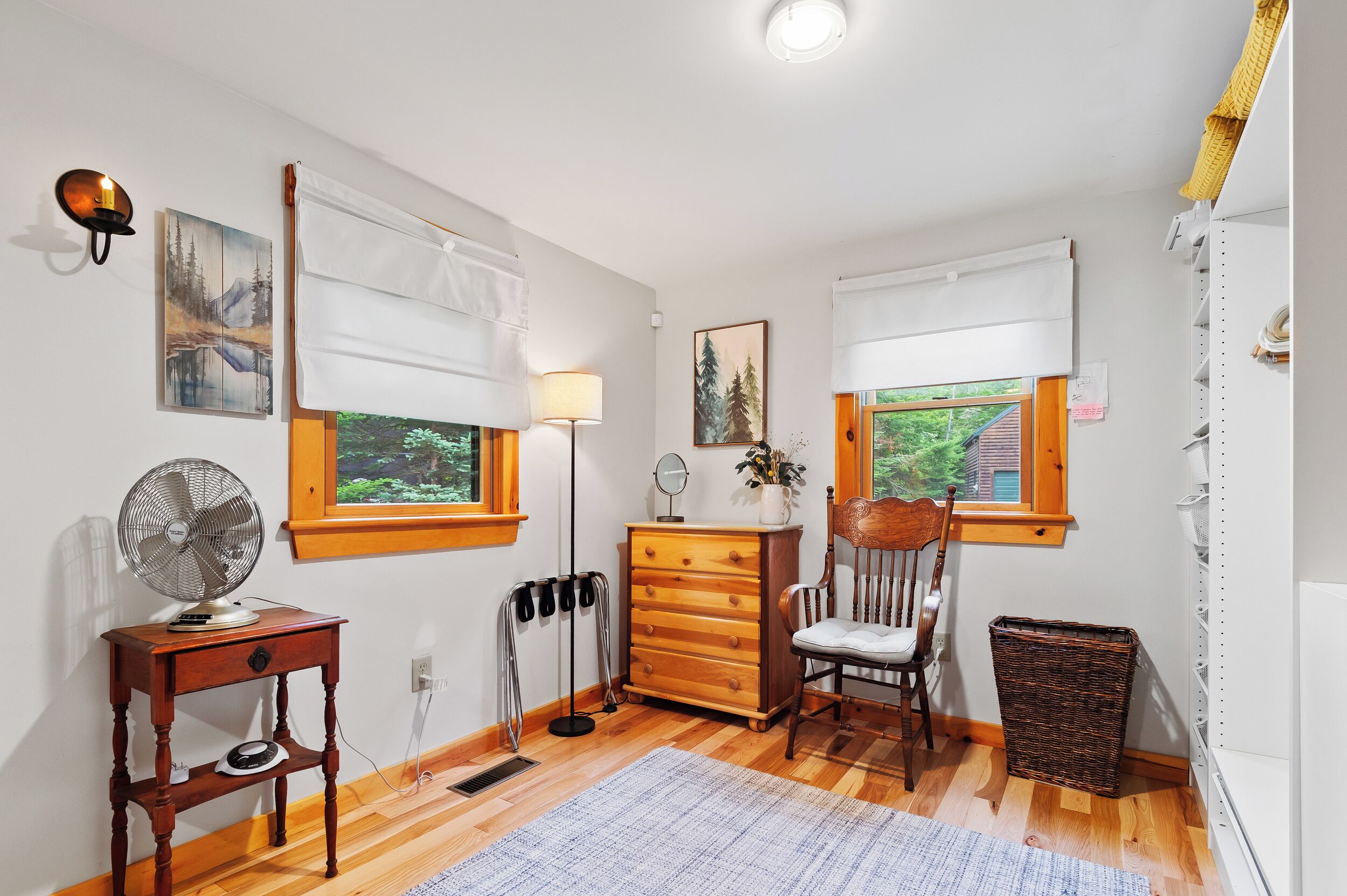 A room with wooden flooring, a wooden dresser, a chair, a standing fan, and a window with a view of greenery.