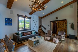 Amplia sala de estar con un sofá gris oscuro, una mesa de centro de madera y un ventanal con vistas a la vegetación. Una escalera de madera conduce a otro nivel, y se observa una puerta de madera rústica.