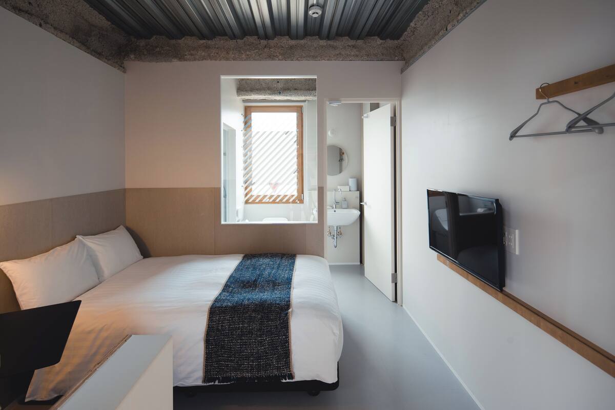 A bedroom with a bed, a TV mounted on the wall, a window, and a bathroom visible through an open door.