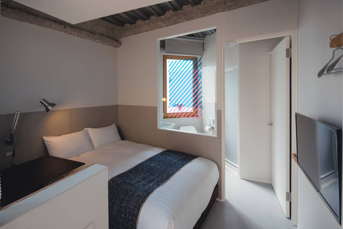A bedroom with a bed, bedside table, lamp, and a window with a view of a building.