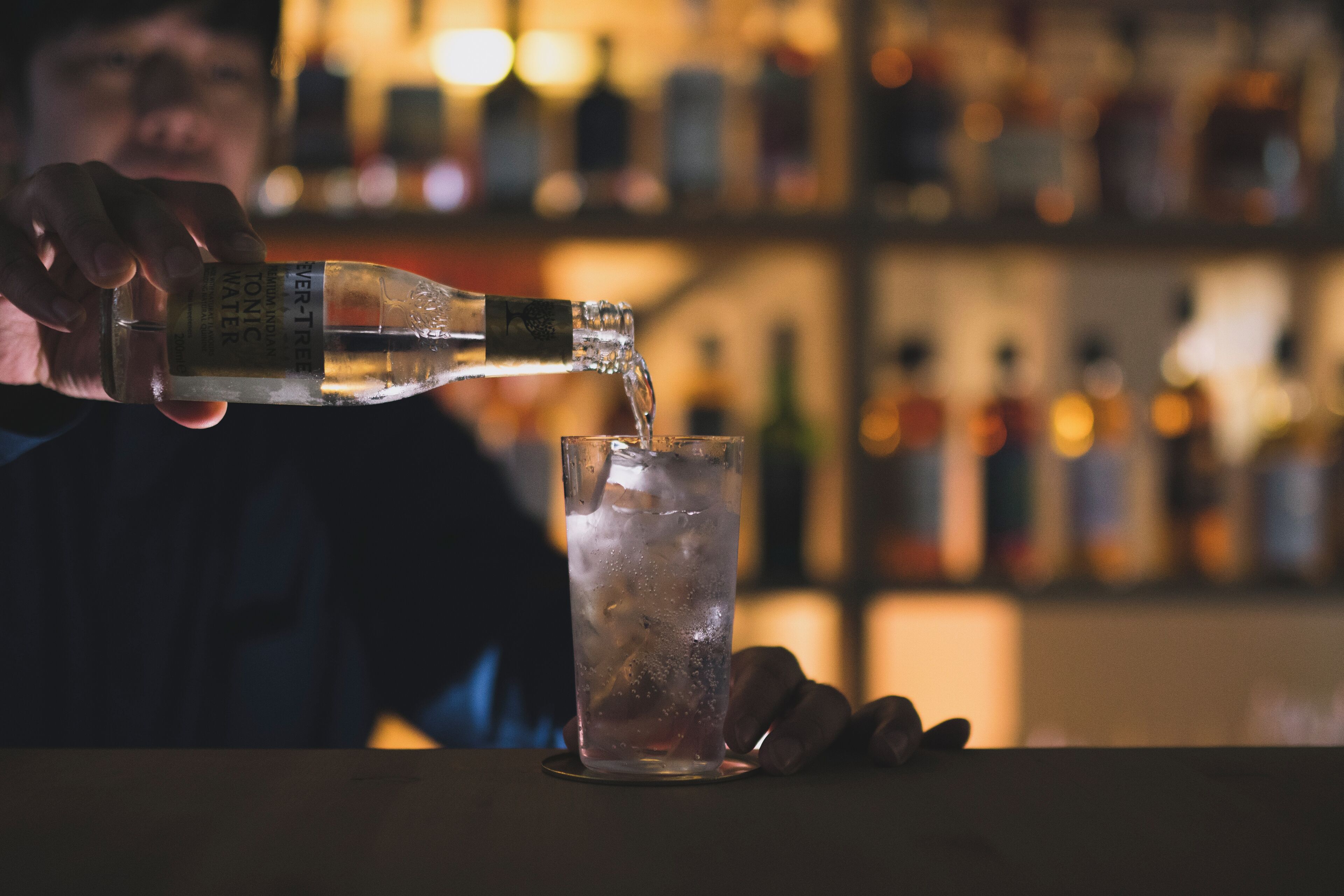 A person pouring a clear liquid from a bottle into a glass with ice cubes.