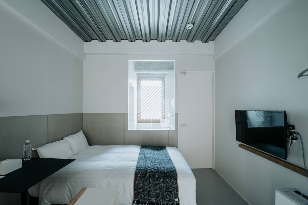 A modern hotel room with a bed, a TV, a desk, and a window.