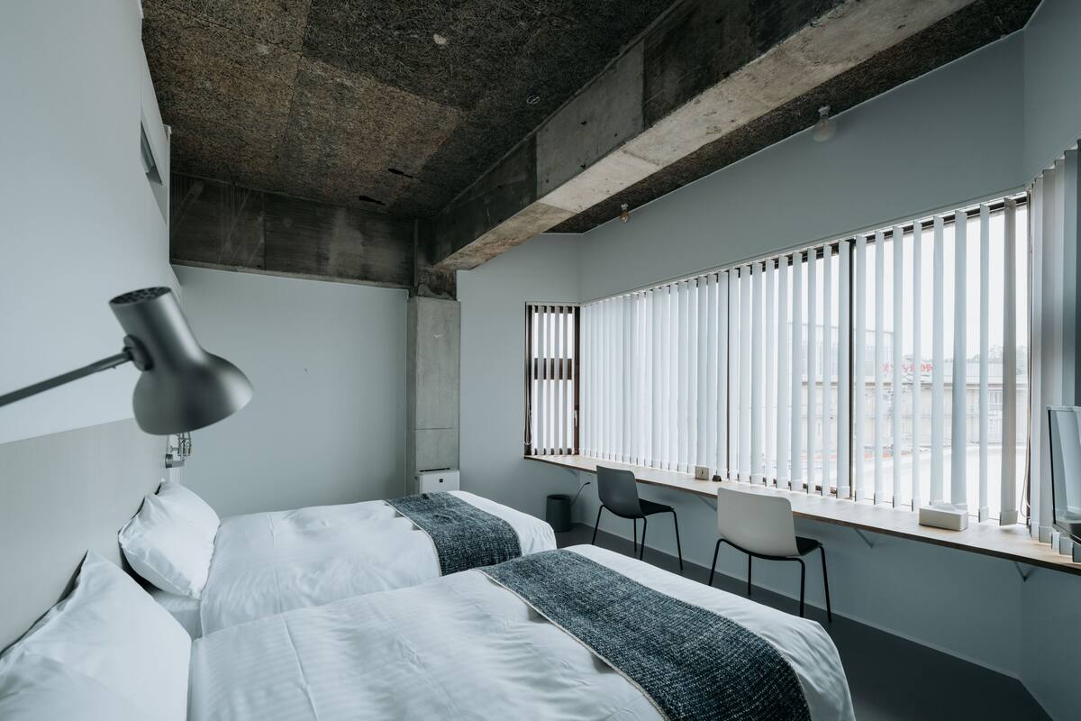 A modern hotel room with two beds, a desk, and a large window with blinds.