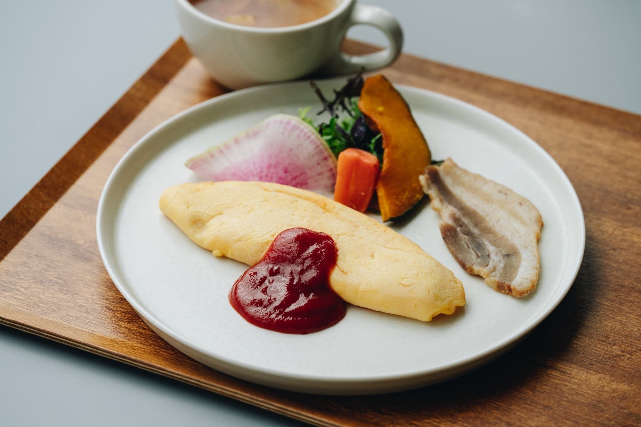 Een bord met een pannenkoek, een stuk vlees, een wortel en een radijs, met een kop koffie op een houten dienblad.