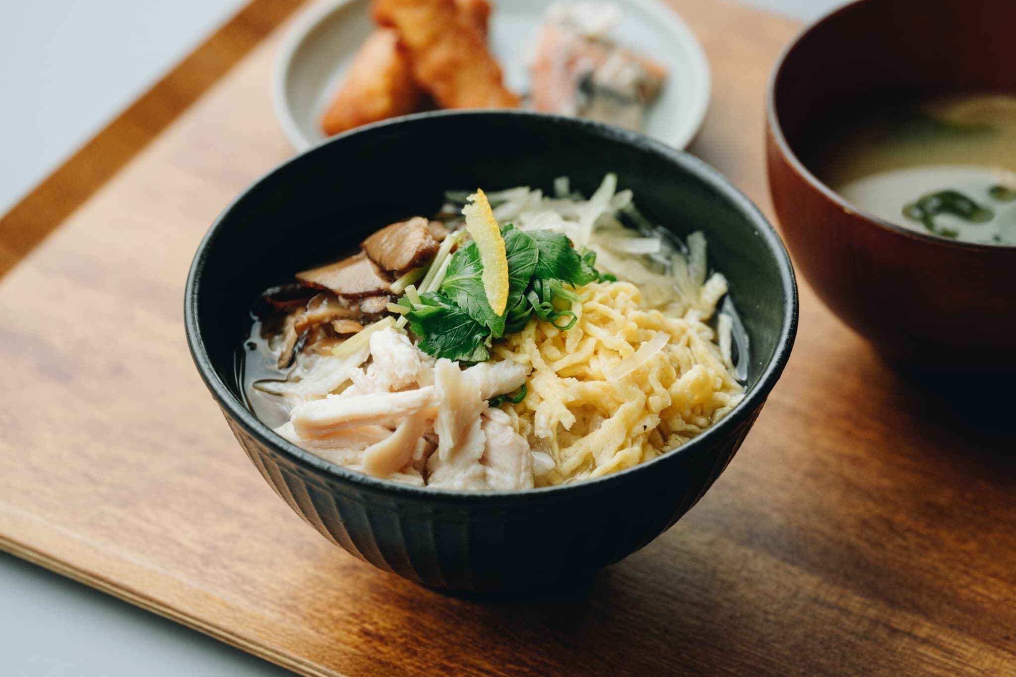 A bowl of noodle soup with chicken and vegetables on a wooden tray.