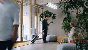 Un moderno vestíbulo de hotel con un amplio ventanal, una zona de estar con sofá y reposapiés, y una planta en maceta.