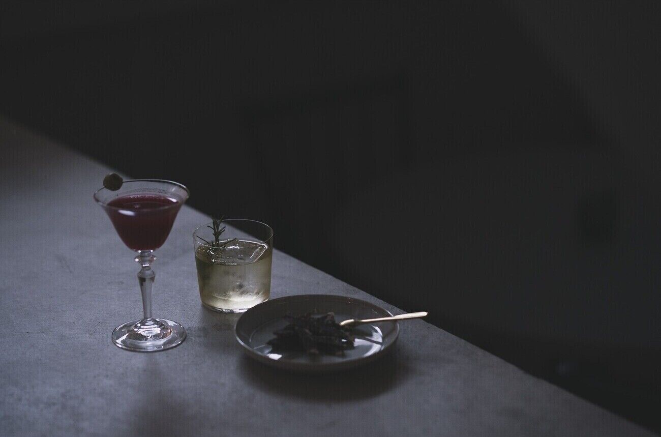 A martini glass with a garnish, a small glass with ice, and a plate with food on a dark surface.