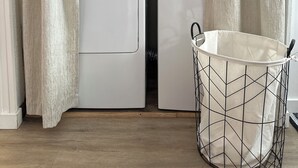 Een wasserette met twee witte voorladers, houten kastjes erboven en een metalen wasmand op de vloer.