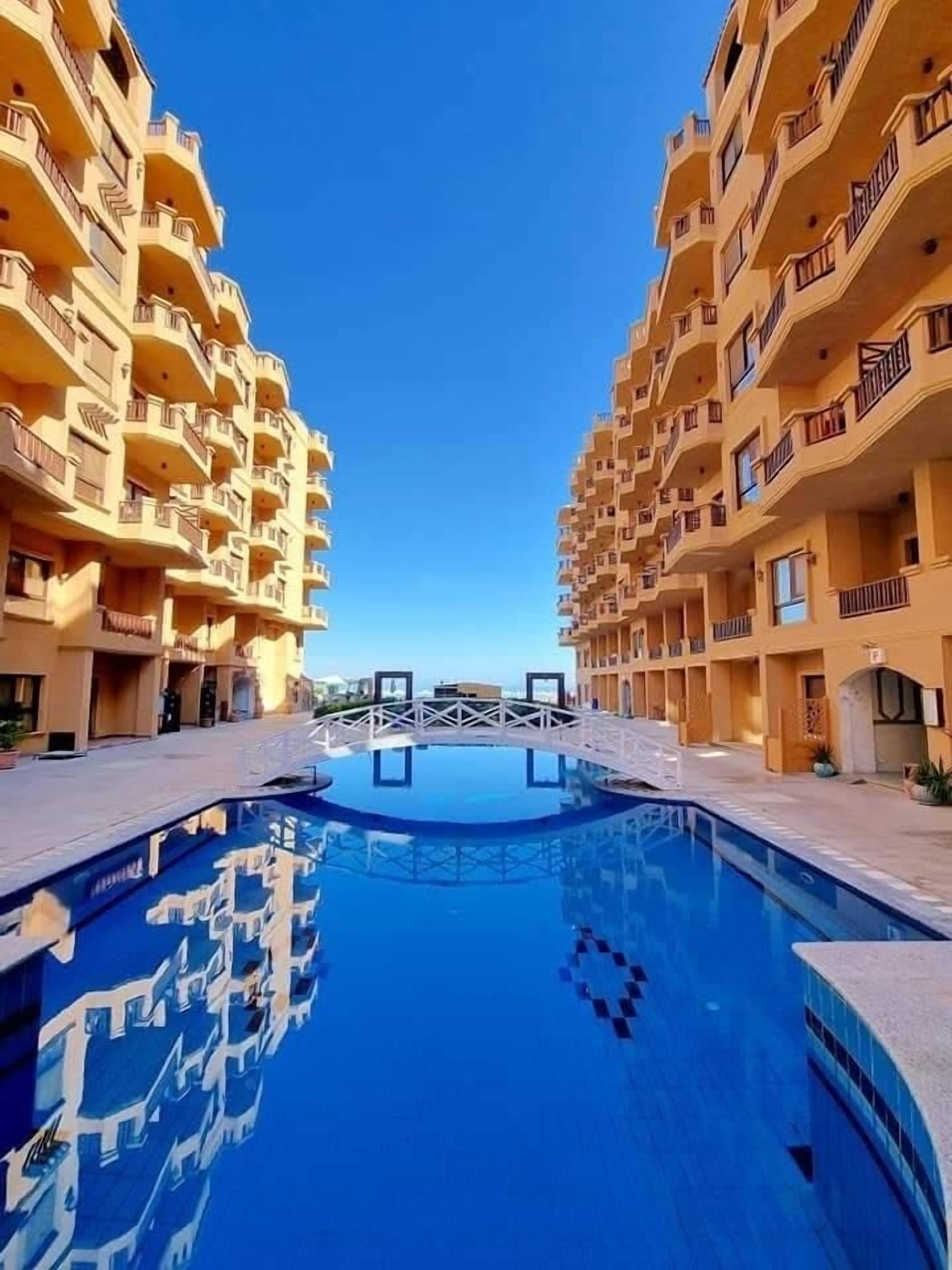 Een hotel met een centraal gelegen zwembad, meerdere balkons en een helderblauwe lucht.