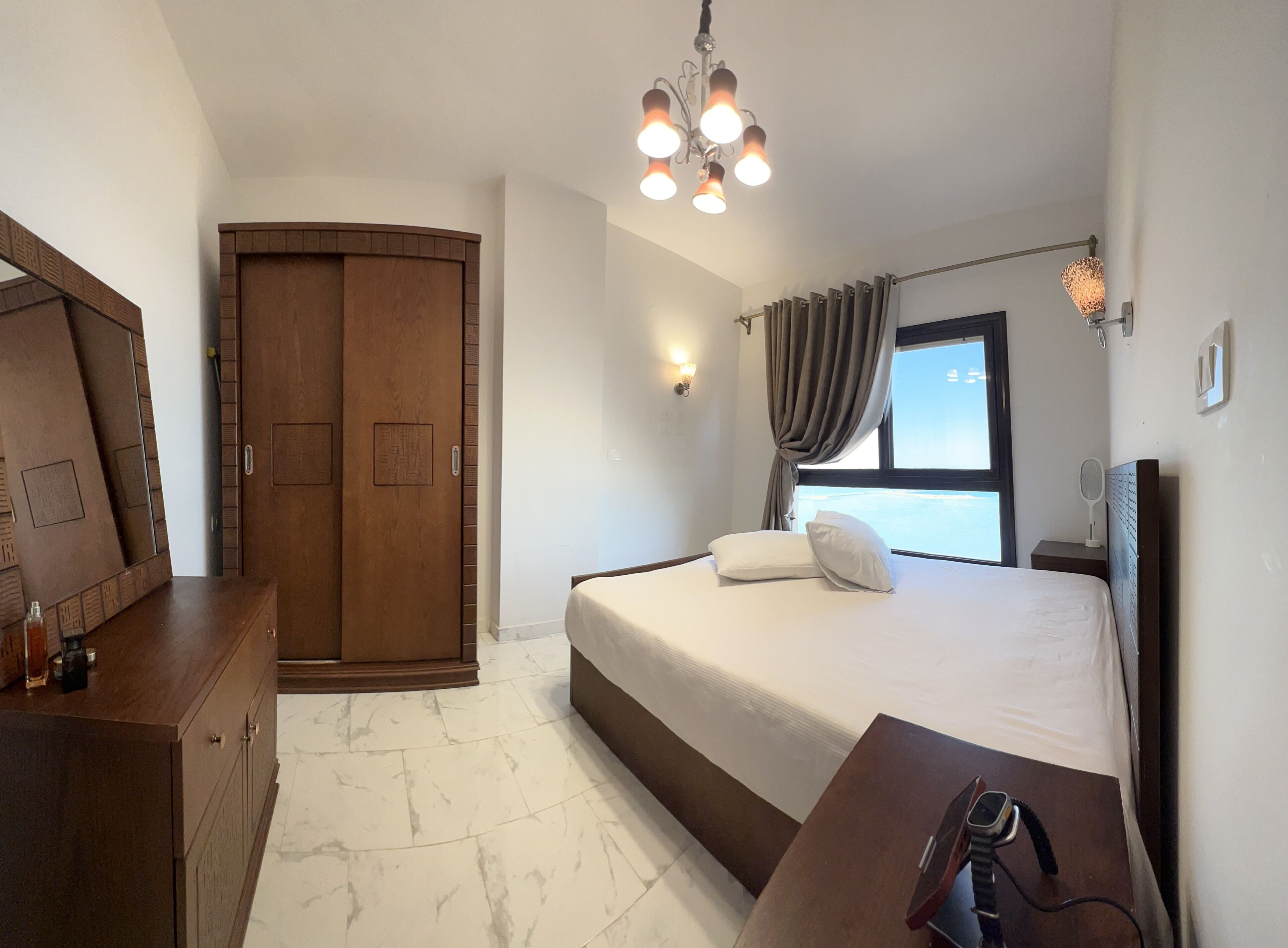 Un dormitorio con cama, un armario de madera, una mesita de noche con un reloj y una ventana con cortinas.