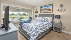 A bedroom with a bed, a desk, a chair, a TV, a window with a view of a golf course, and a painting of a beach scene. - Legend Villas of Bay Point 4261 (Panama City Beach)