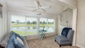 A room with a view of a golf course through large windows, a ceiling fan, a blue chair, a gray couch, and a small table with a decorative item. - Legend Villas of Bay Point 4261 (Panama City Beach)
