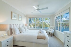 Une chambre dotée d’un grand lit, d’un ventilateur de plafond, d’une chaise et offrant une vue sur une étendue d’eau grâce à de grandes fenêtres.