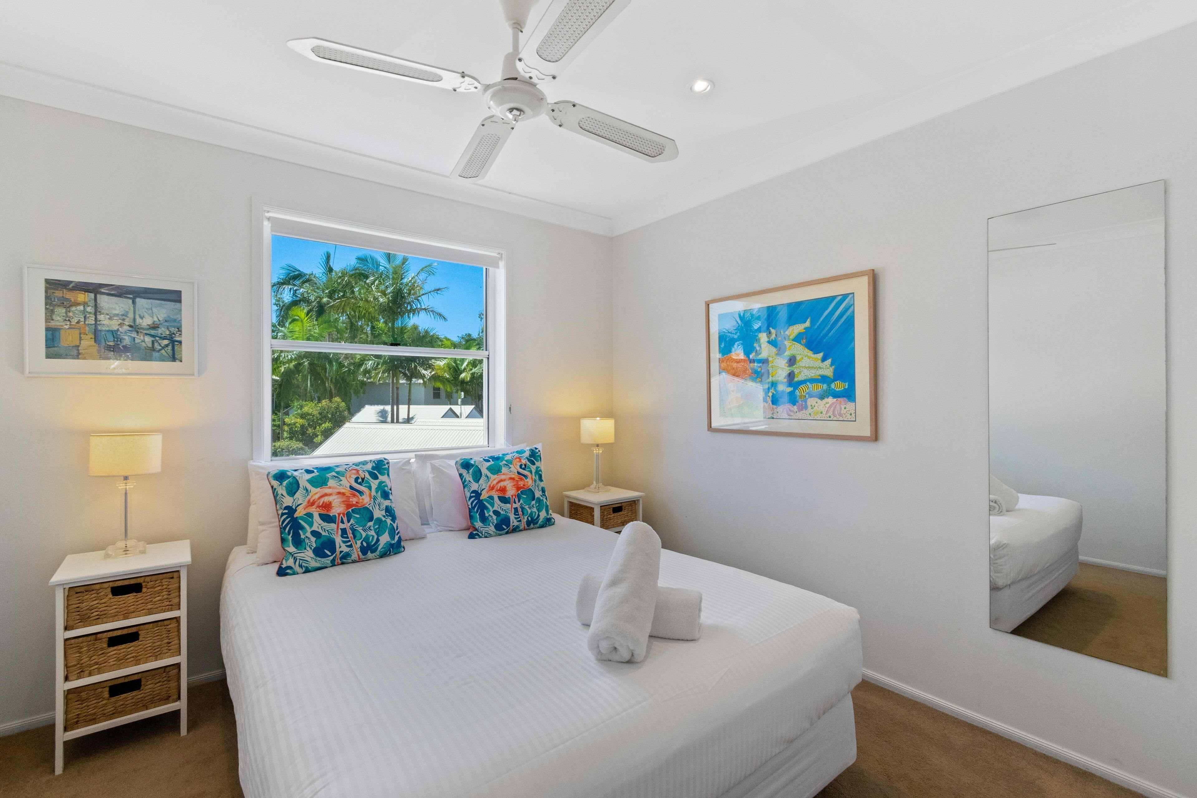 A bedroom with a large bed, a ceiling fan, a window with a view of palm trees, and two bedside tables with lamps.