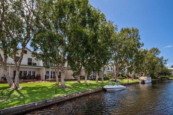 A riverside view with a boat docked on the water, trees lining the path, and a building with a lawn and outdoor seating area.