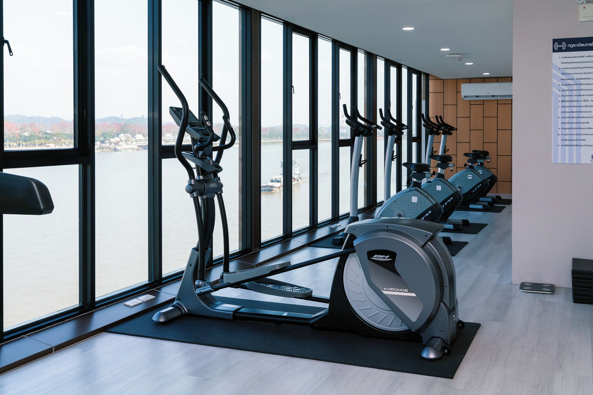A gym with stationary bikes and a rowing machine, large windows with a view of a body of water and buildings, and a wall-mounted information board.