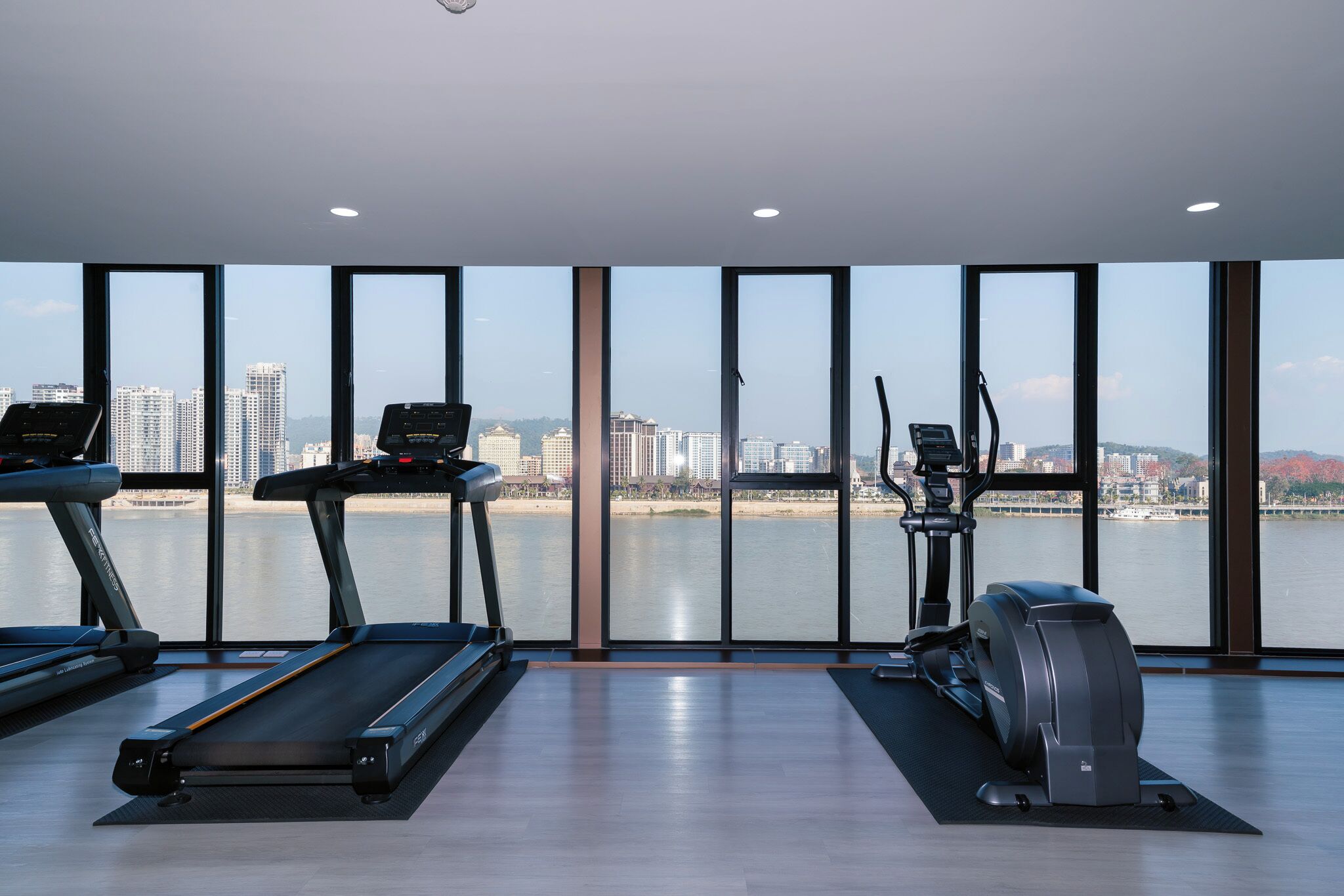 A gym with treadmills, an elliptical, and a rowing machine, overlooking a cityscape and a body of water.