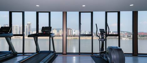 A gym with treadmills, an elliptical, and a rowing machine, overlooking a cityscape and a body of water.
