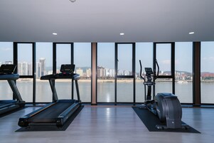 Un gimnasio con cintas de correr, elíptica y máquina de remo, con vistas a un paisaje urbano y una masa de agua.