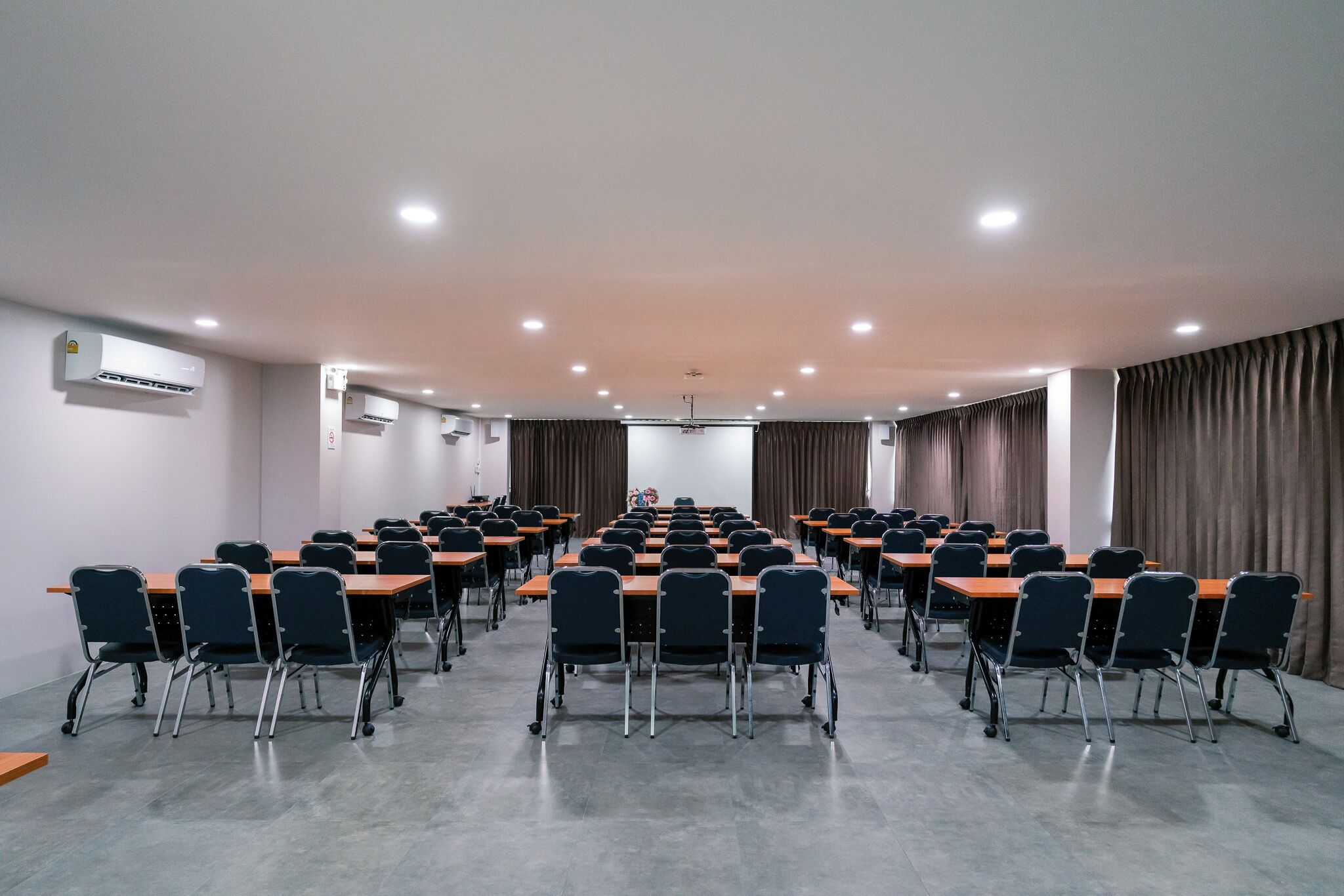 Un salón de conferencias con mesas y sillas dispuestas en filas, unidades de aire acondicionado y una pantalla de proyector.