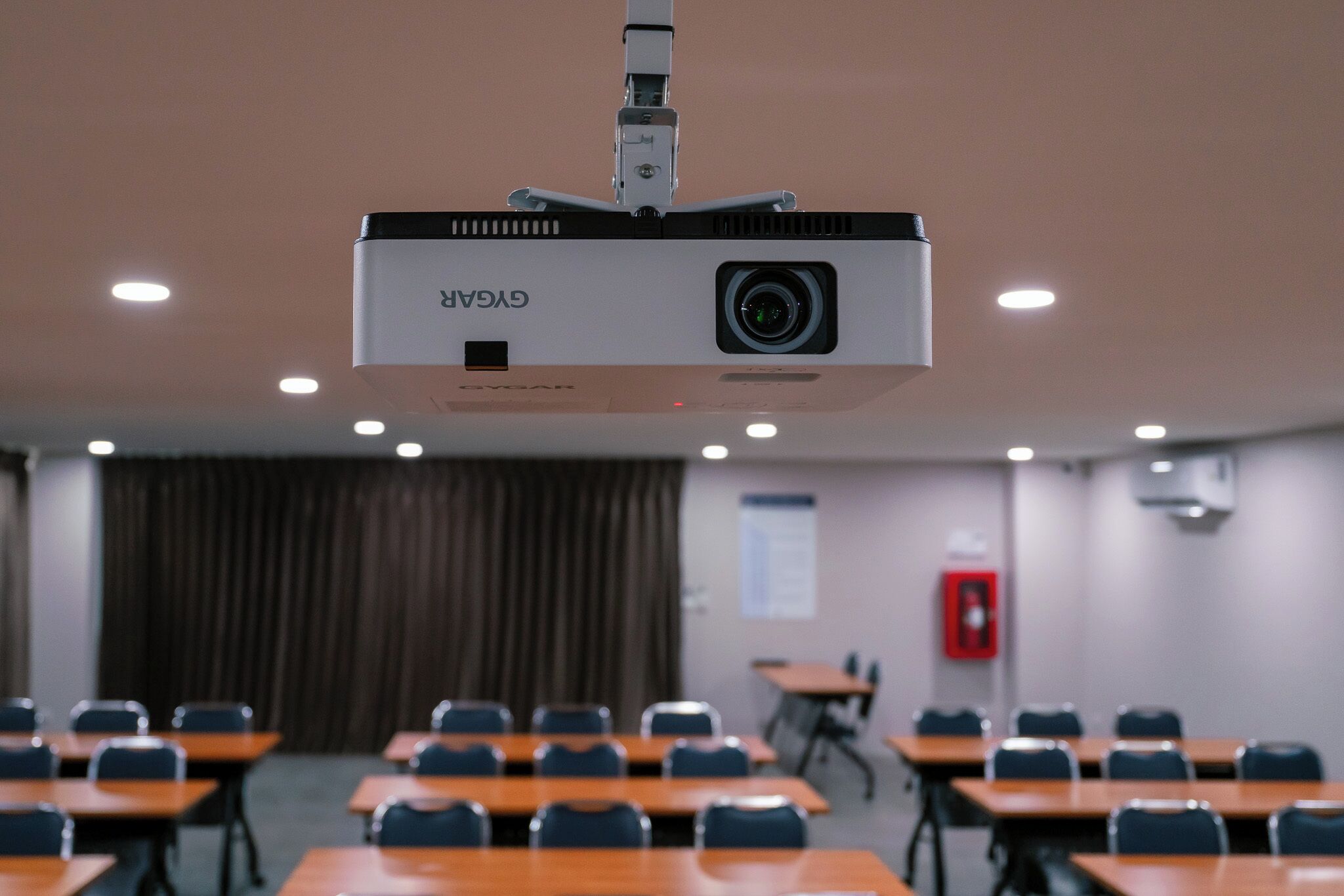 A conference room with a ceiling-mounted projector, tables, chairs, and a fire extinguisher on the wall.