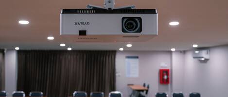 A conference room with a ceiling-mounted projector, tables, chairs, and a fire extinguisher on the wall.