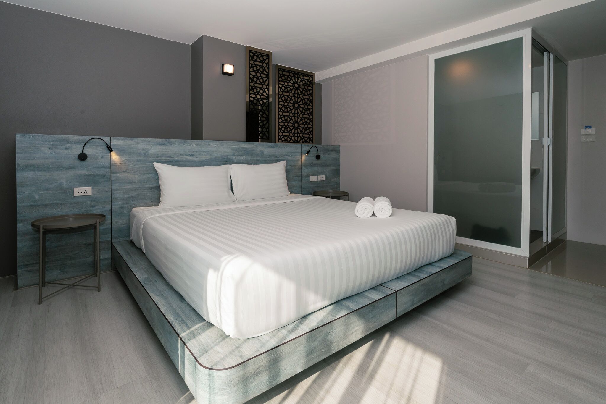 A modern hotel room with a large bed, bedside tables, a glass shower enclosure, and a decorative wall panel.