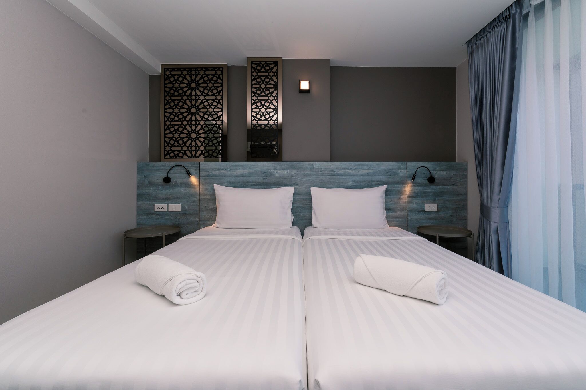 Una cama doble con dos almohadas y dos toallas enrolladas, una mesita de noche con una lámpara y un panel decorativo sobre la cama.