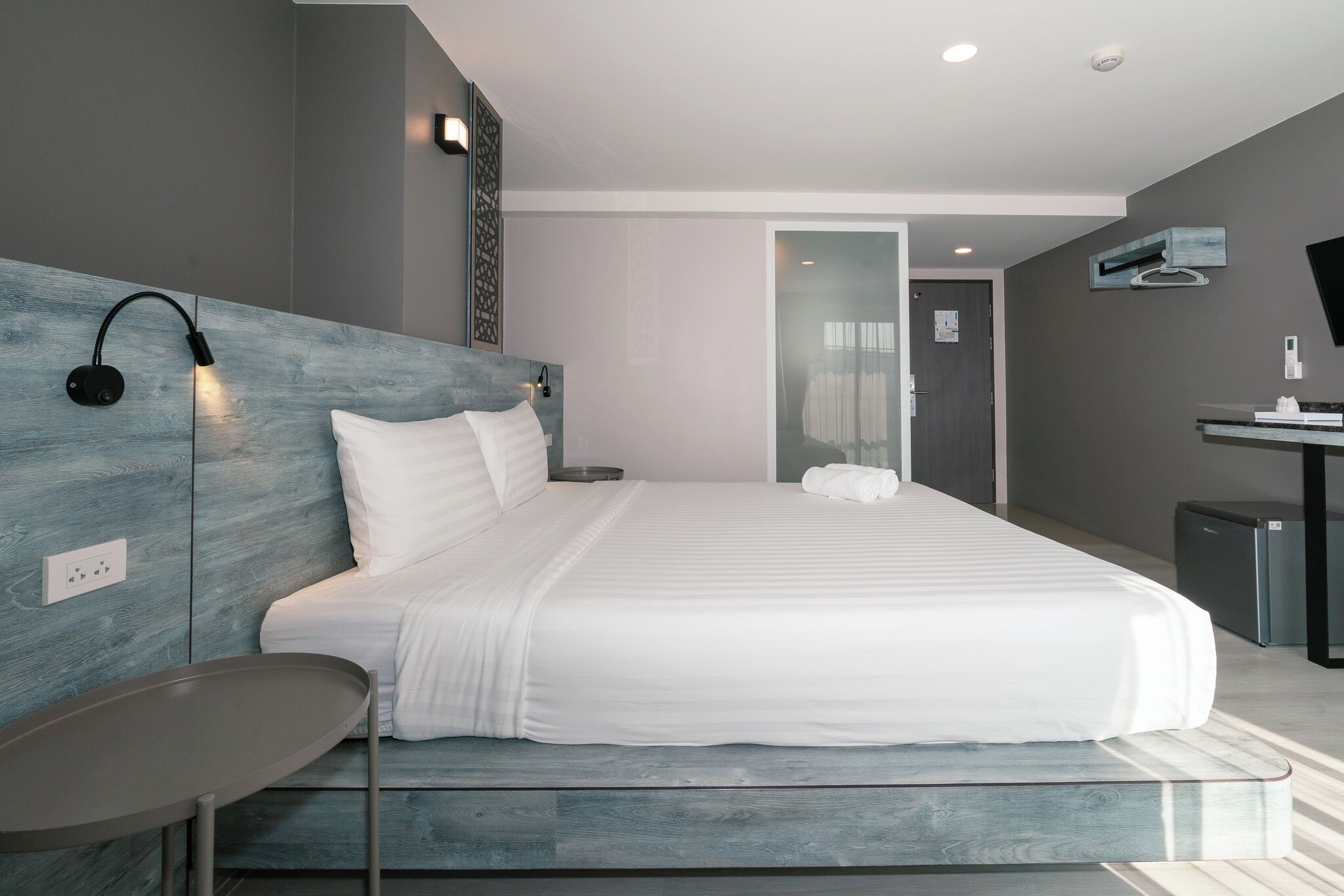 A modern hotel room with a large bed, bedside table, and a desk with a chair.